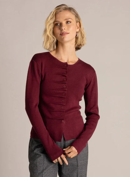 A woman modeling a long-sleeve knit top in deep burgundy with a buttoned front and gathered detail at the chest, paired with gray textured pants, against a plain light gray studio background.