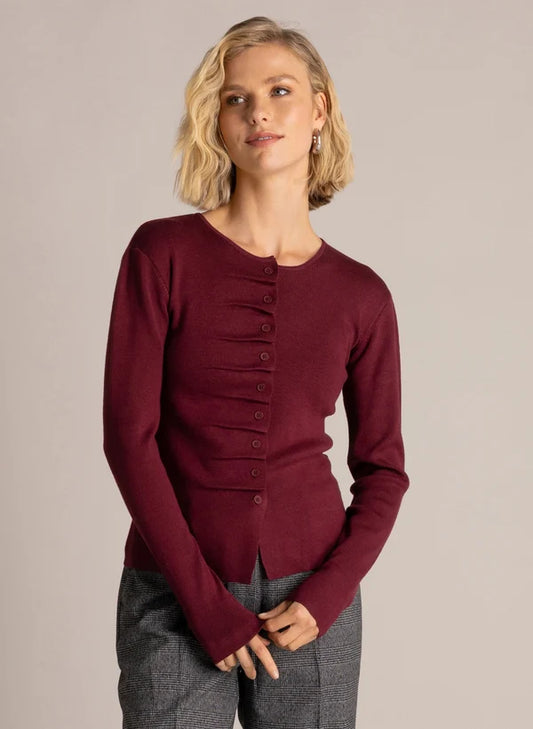 A woman modeling a long-sleeve knit top in deep burgundy with a buttoned front and gathered detail at the chest, paired with gray textured pants, against a plain light gray studio background.