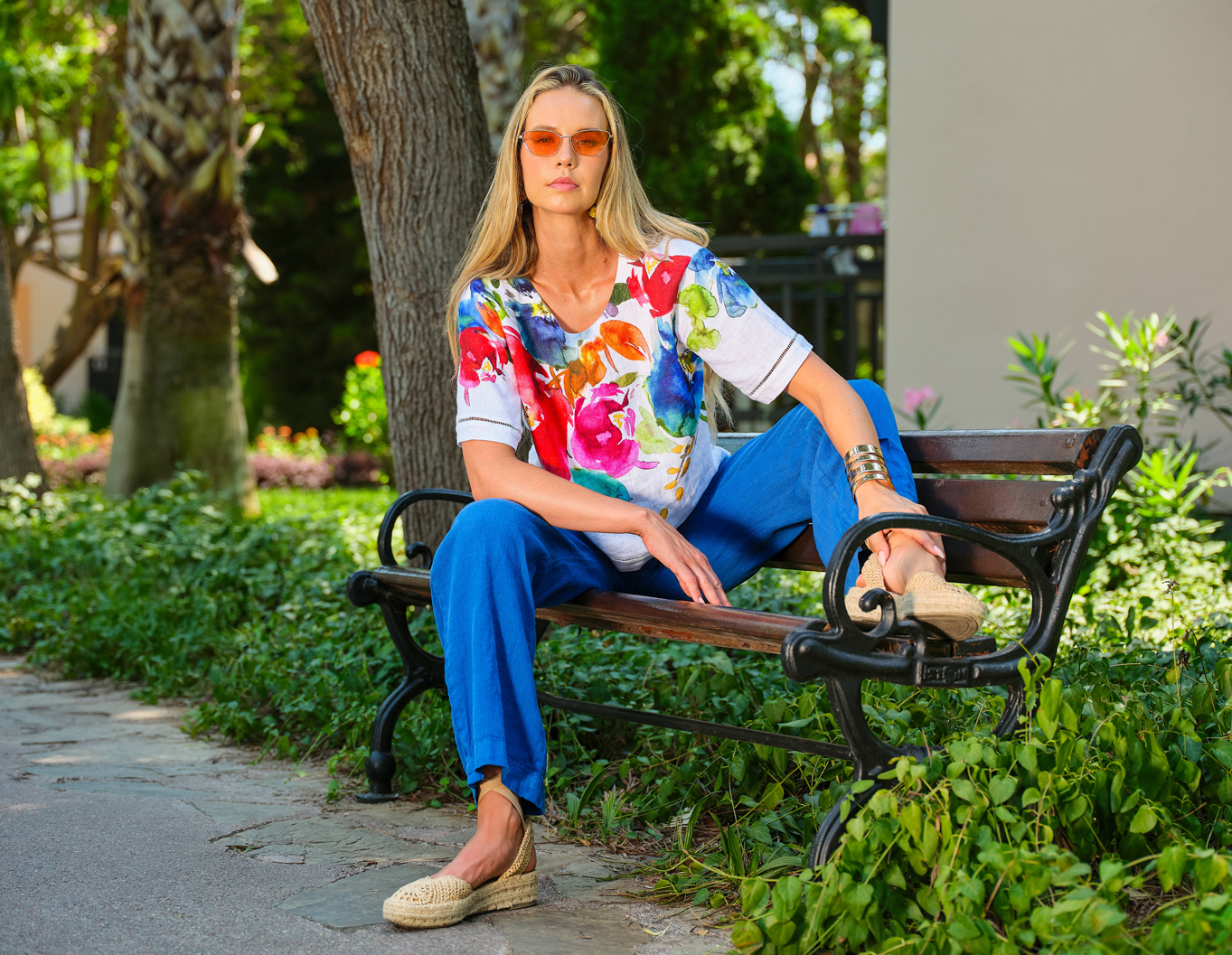 Woman sitting on a bench wearing a colorful floral shirt and blue pants in a park setting.