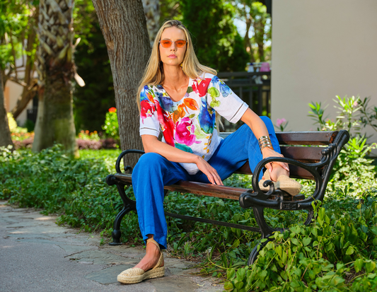 Woman sitting on a bench wearing a colorful floral shirt and blue pants in a park setting.