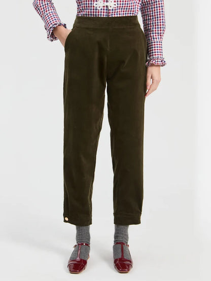 A model is shown from the waist down wearing dark olive green corduroy trousers, a red and white gingham shirt, gray socks, and red Mary Jane shoes against a plain white studio background.