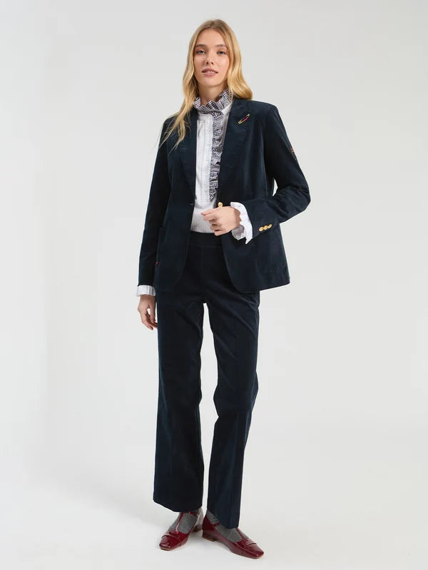 A woman modeling a dark navy blue two-piece velvet  suit with a patterned white shirt and dark red loafers, standing against a light gray studio background.
