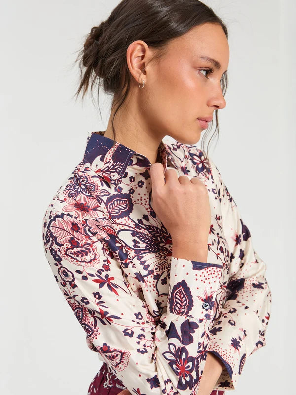 A woman in profile wearing a long-sleeve button-up shirt / Blouse with a dense floral and paisley print in navy blue, burgundy, and cream, set against a plain light gray studio background.