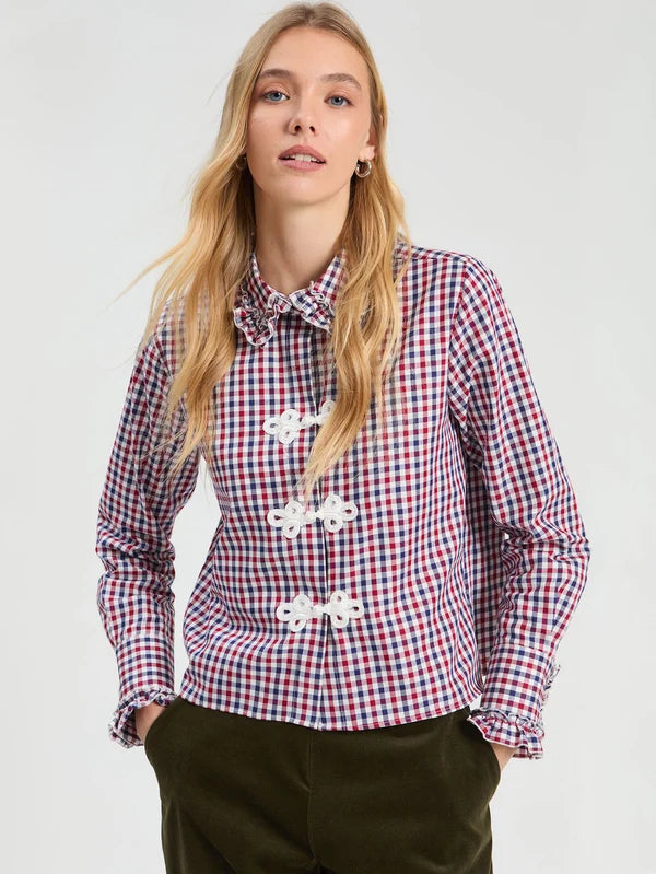 A woman modeling a red, white, and blue gingham long-sleeve blouse with white decorative frog closures and a matching bow tie collar, paired with dark olive pants against a light gray studio background.