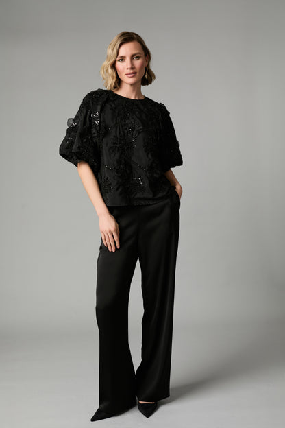 A woman modeling a black embellished short-sleeve top with puffed sleeves and black riley wide-leg trousers, standing 