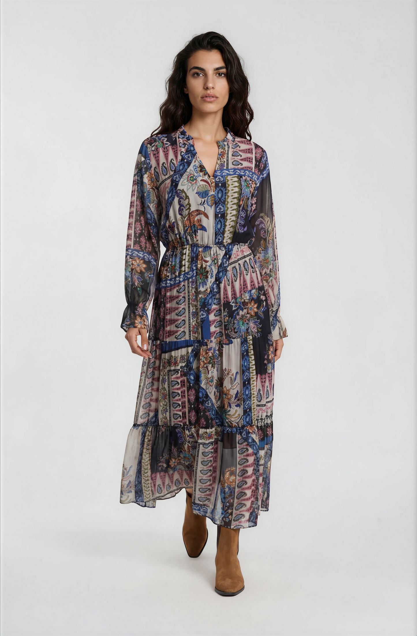 A woman modeling a long-sleeved midi dress with a multicolor abstract and geometric print in shades of blue, pink, purple, and beige, paired with brown ankle boots, against a plain light gray studio background.