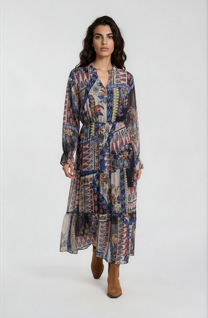 A woman modeling a long-sleeved midi dress with a multicolor abstract and geometric print in shades of blue, pink, purple, and beige, paired with brown ankle boots, against a plain light gray studio background.