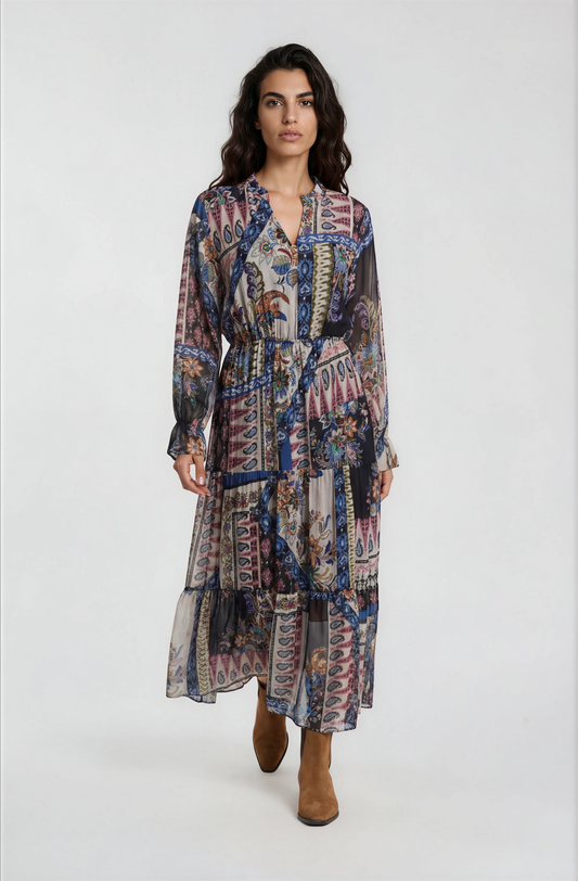 A woman modeling a long-sleeved midi dress with a multicolor abstract and geometric print in shades of blue, pink, purple, and beige, paired with brown ankle boots, against a plain light gray studio background.