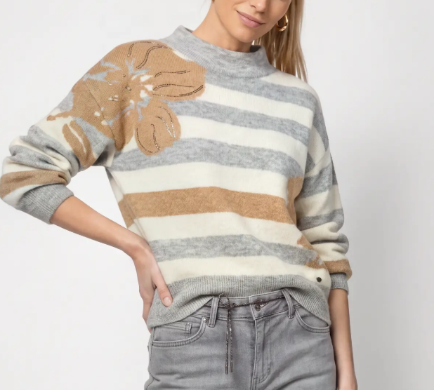 A woman wearing a mock-neck knit sweater with horizontal gray and cream stripes, a large tan floral applique on the left chest, and gray jeans, photographed against a plain light background.