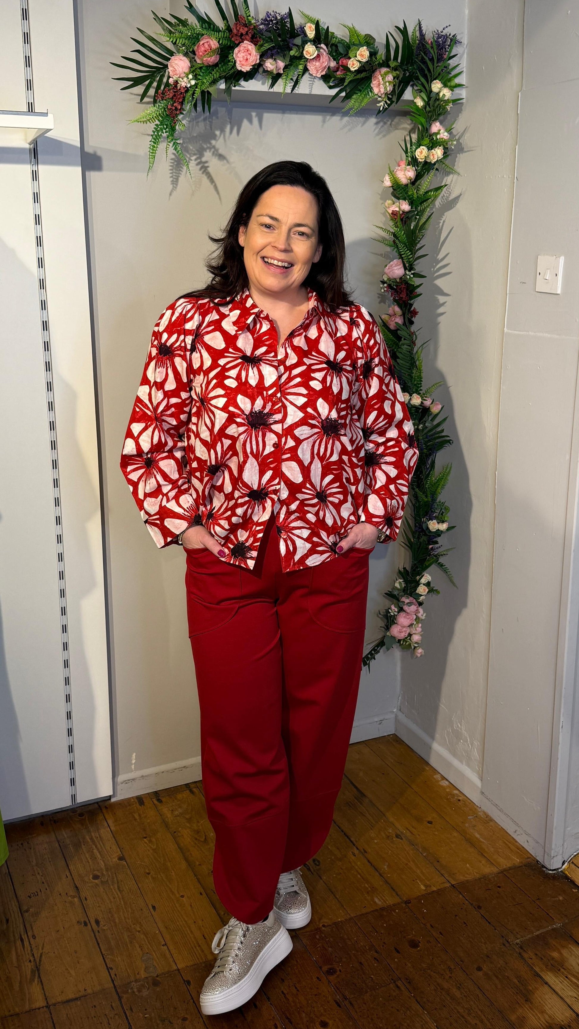 Person wearing a red and white patterned top and red pants standing in front of a floral arch.