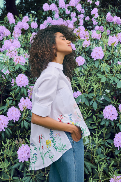 Women’s white embroidered blouse with multicolour floral hem detail, short sleeves and stand collar, styled with denim jeans.