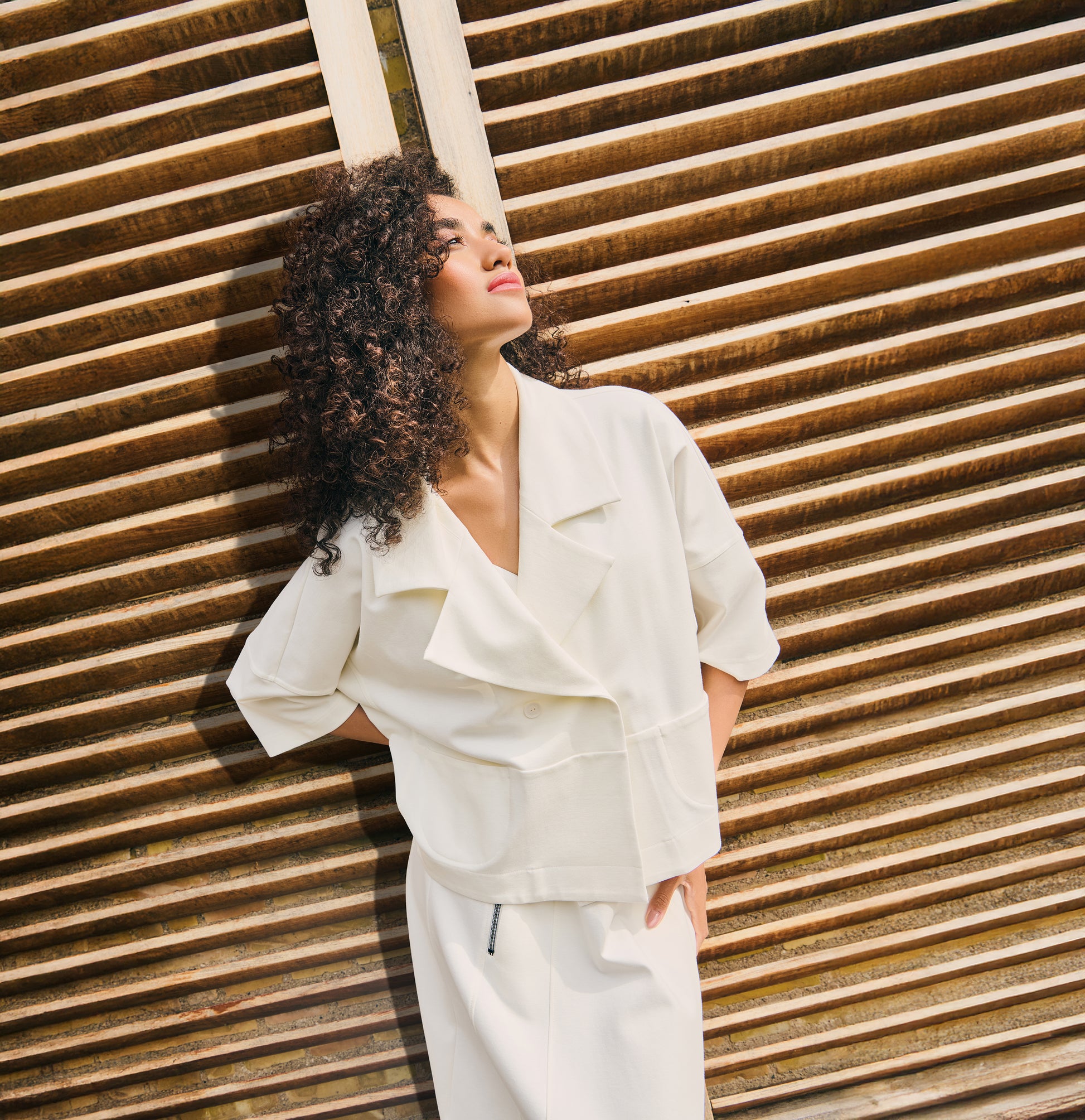 Model wearing an oversized white jacket top with wide lapel collar and short sleeves, styled in a relaxed fit against a wooden slatted background.