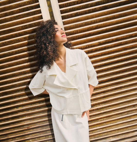 Model wearing an oversized white jacket top with wide lapel collar and short sleeves, styled in a relaxed fit against a wooden slatted background.
