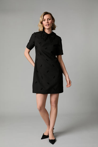 A woman modeling a short-sleeved black shift dress with a decorative ruffled collar and subtle tonal embroidered details, paired with black pointed-toe heels against a neutral gray studio background.