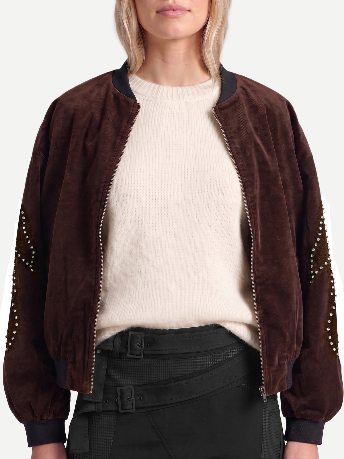 A model wearing a dark brown velvet bomber jacket with studded details on the sleeves, layered over a cream knit sweater and black pants.