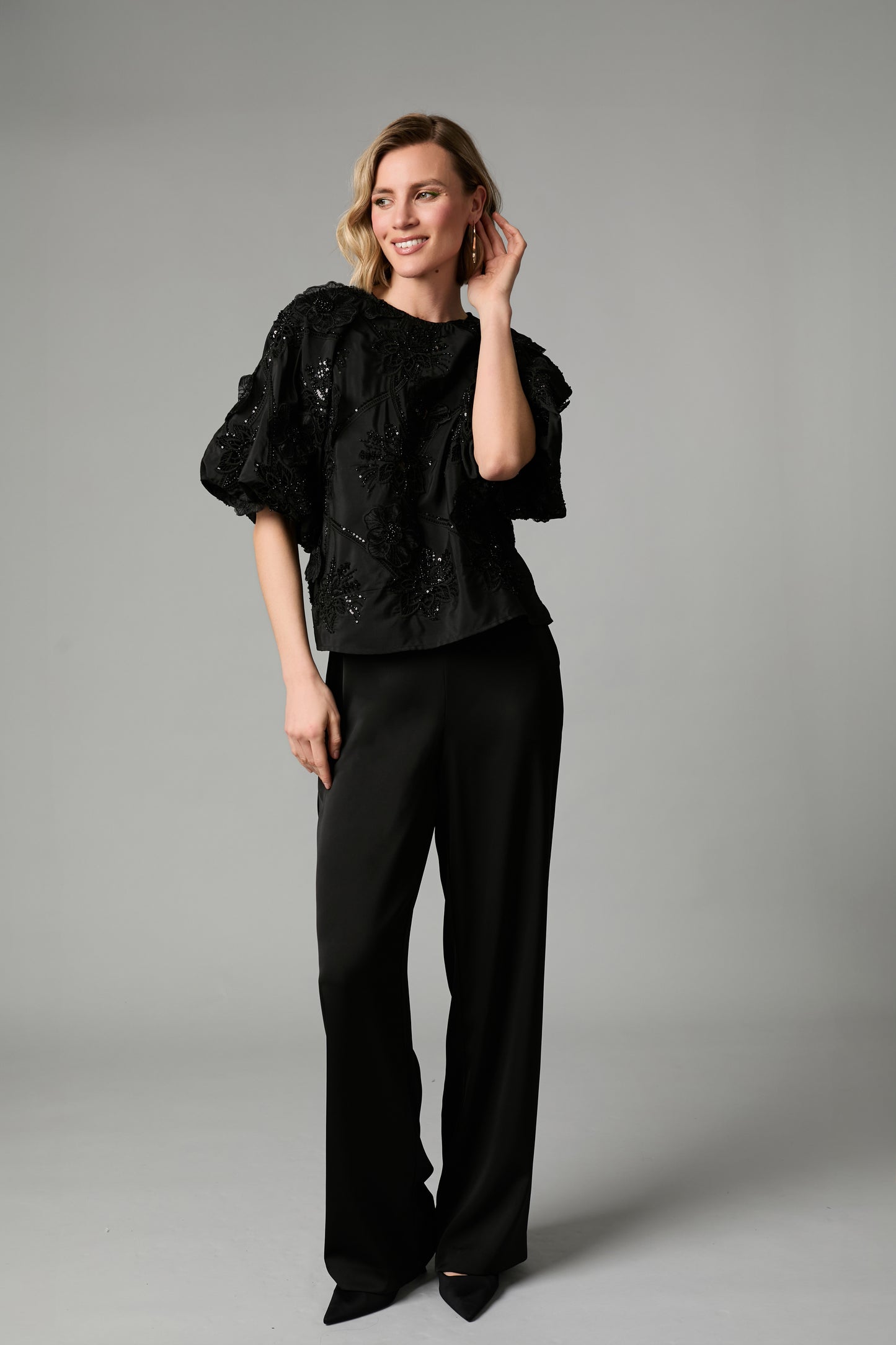 A woman modeling a black ruffled short-sleeve top with sequined detailing and black wide-leg trousers, standing against a neutral gray studio background.