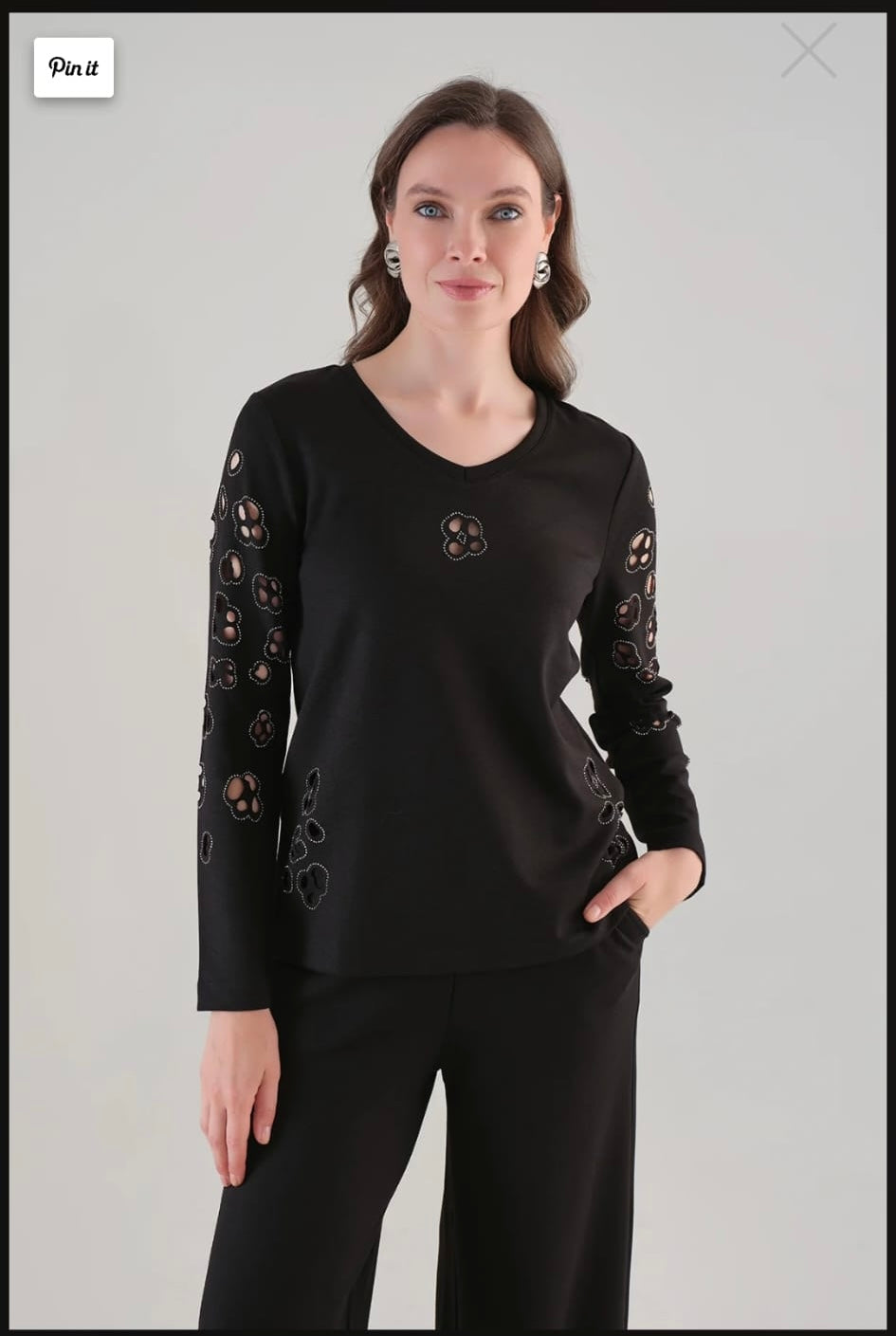 A woman modeling a black long-sleeve top with decorative cutout lace details on the sleeves and a small circular embroidered motif on the chest, paired with black pants, against a light gray studio background.