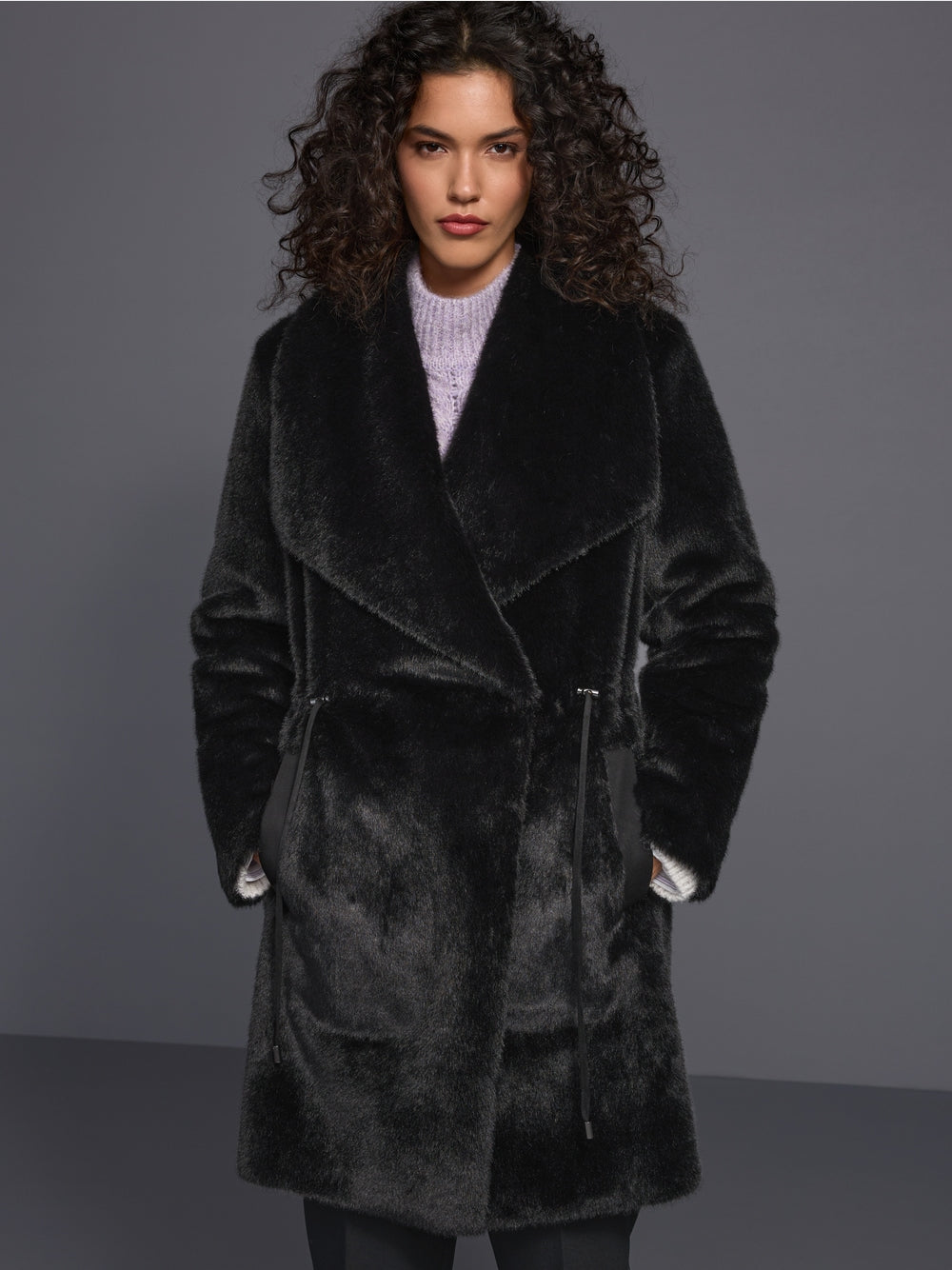 A model with curly dark hair wearing a plush black faux fur wrap coat over a light purple knit sweater, standing against a dark gray studio background.