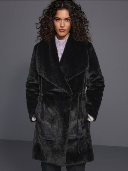 A model with curly dark hair wearing a plush black faux fur wrap coat over a light purple knit sweater, standing against a dark gray studio background.