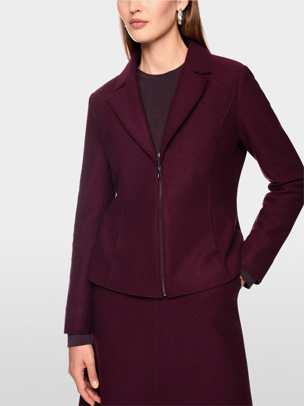 A woman wearing a structured, deep burgundy zip-front jacket with notched lapels and a matching skirt, photographed against a clean light gray studio background.