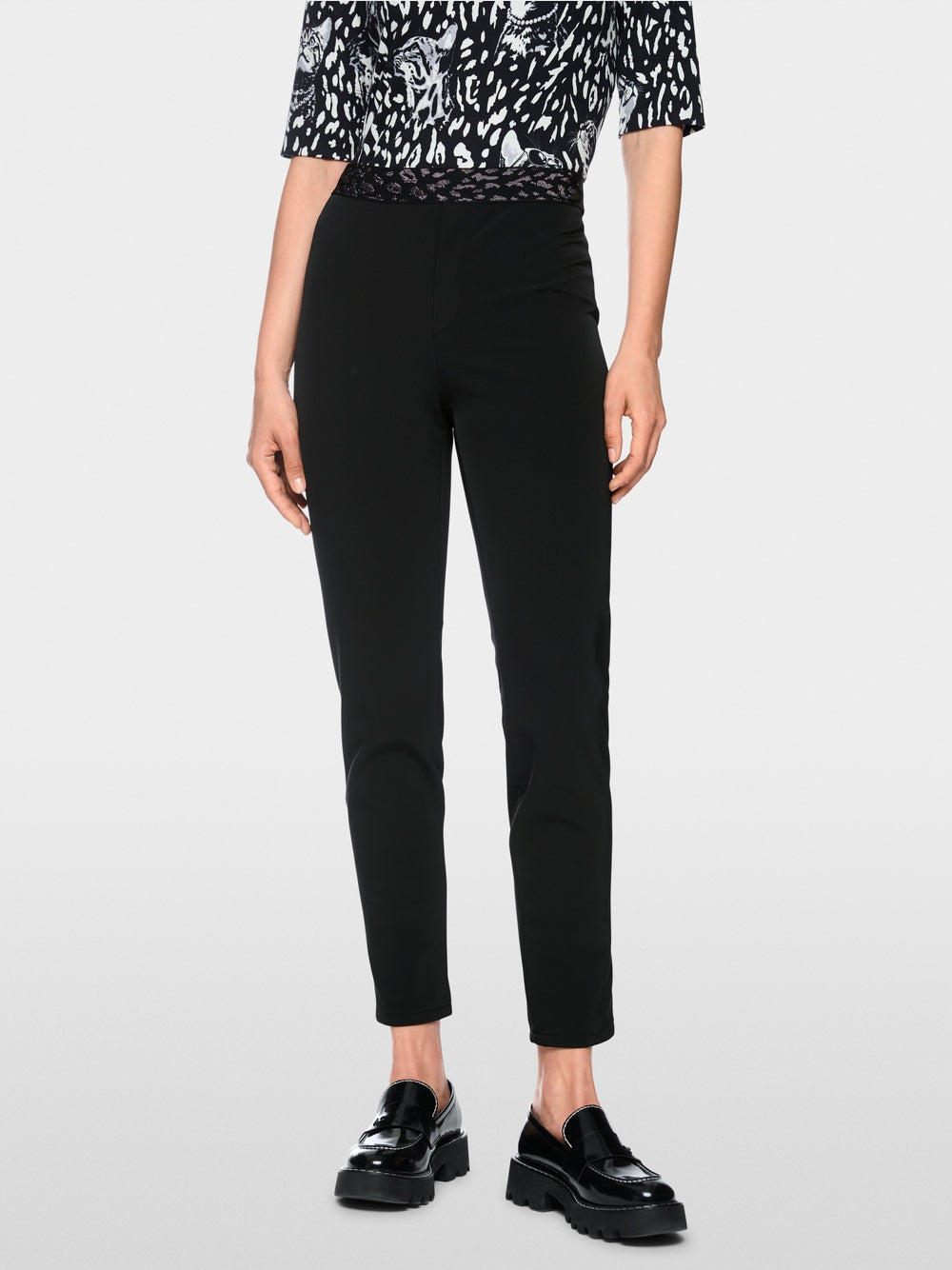 A model is shown from the chest down wearing slim black cropped trousers, a black and white patterned top, and black leather loafers against a light gray studio background.