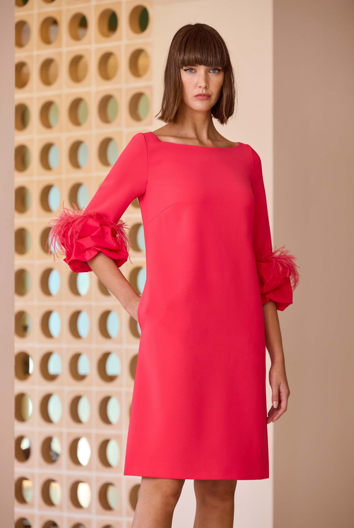 Model wearing a bright pink shift dress with boat neckline and feather trim three-quarter sleeves, styled for a modern occasion look