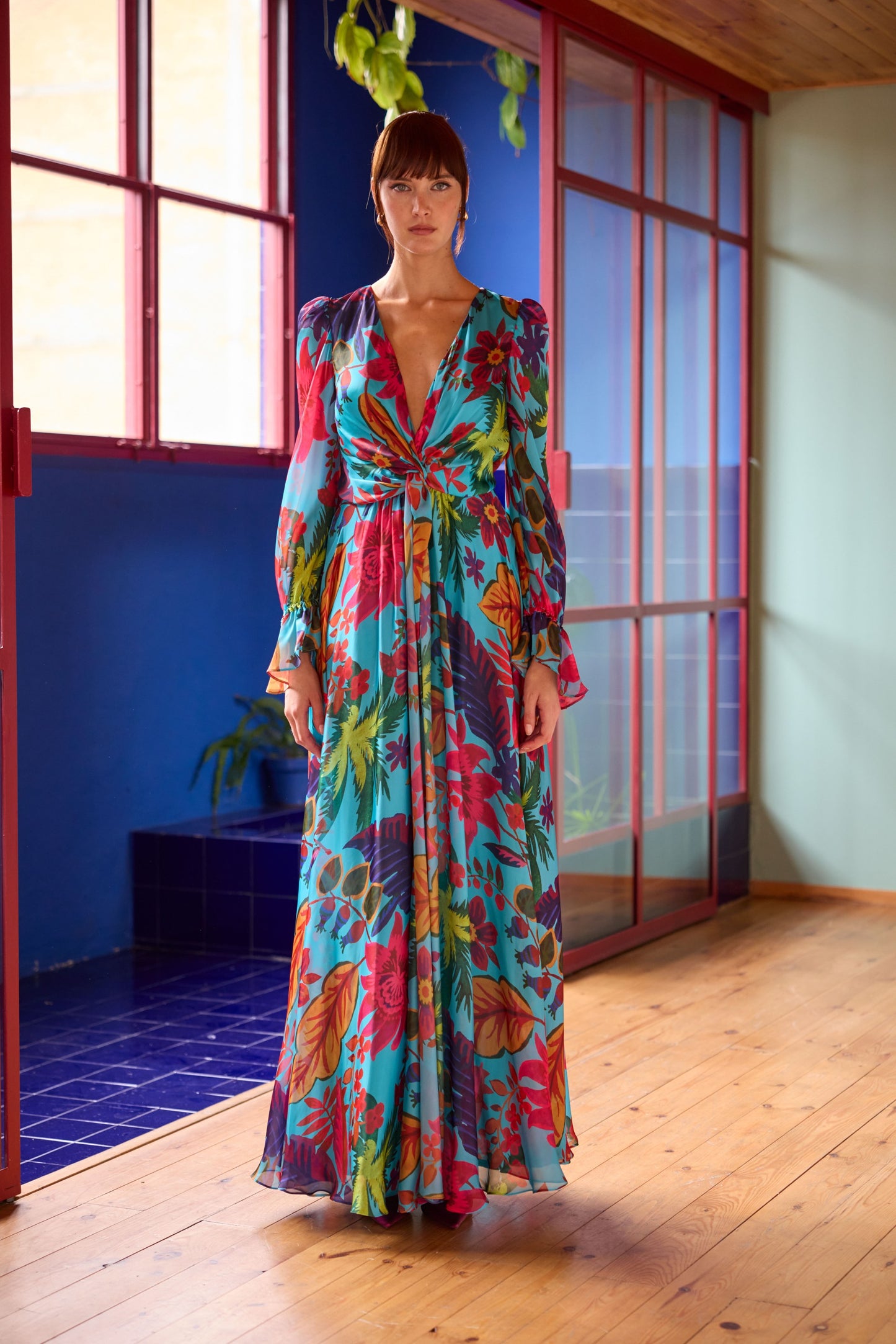 Model wearing a turquoise maxi dress with a multicolour tropical floral print, featuring a deep V-neckline, long sheer sleeves, and a flowing full-length skirt, styled indoors with colourful architectural details