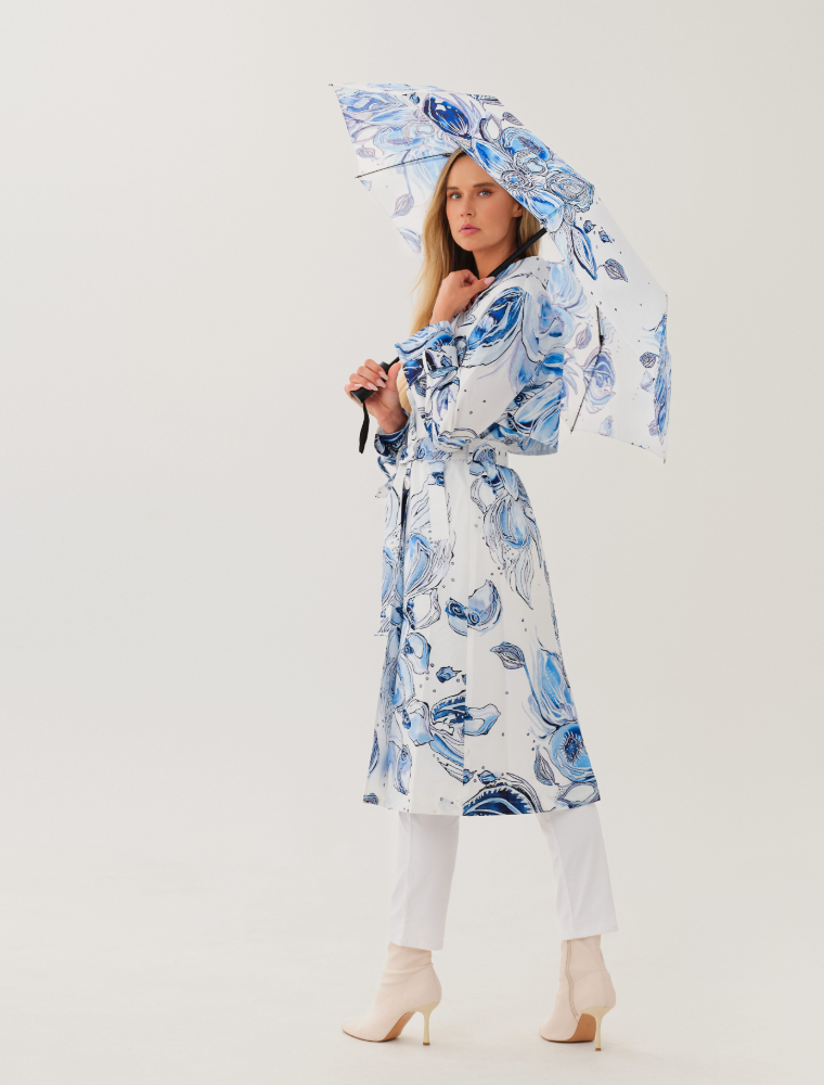 Model wearing a white Dolcezza trench coat featuring blue abstract botanical artwork by Amy Tiernan, belted at the waist and paired with white trousers and cream ankle boots, holding a matching printed umbrella.
