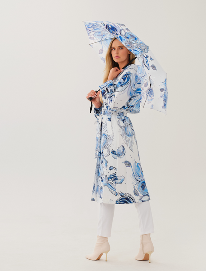 Model wearing a white Dolcezza trench coat featuring blue abstract botanical artwork by Amy Tiernan, belted at the waist and paired with white trousers and cream ankle boots, holding a matching printed umbrella.