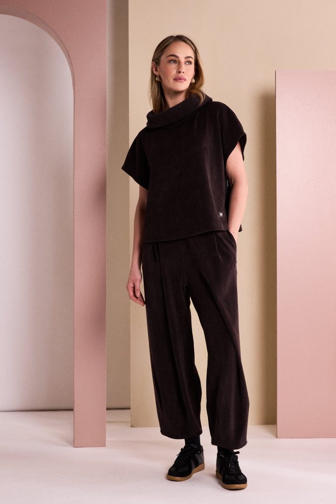 A woman modeling a dark brown short-sleeve mock-neck top and matching wide-leg trousers, standing in a minimalist studio with pink and beige geometric walls.