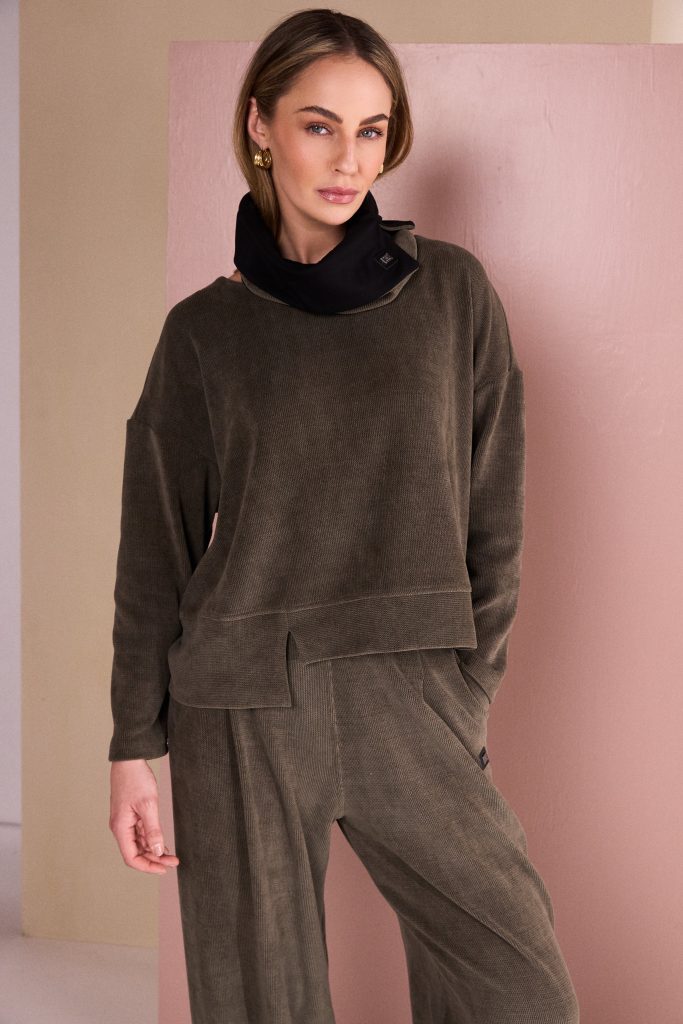 A woman wearing a matching dark brown corduroy lounge set consisting of a cropped long-sleeve top with a black cowl neck and wide-leg pants, posing against a pink and beige wall.
