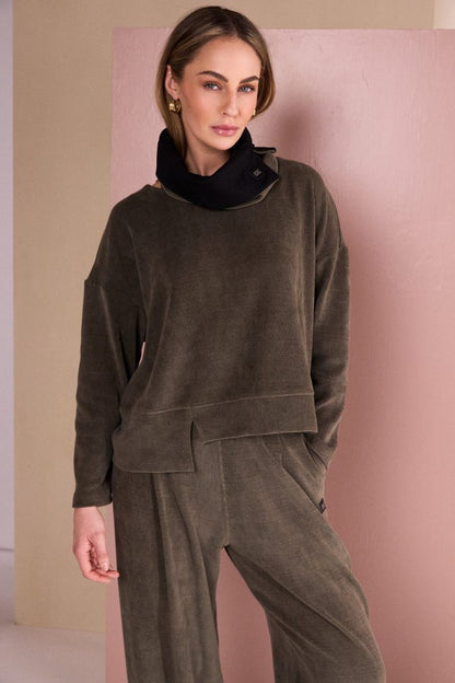 A woman wearing a matching dark brown corduroy lounge set consisting of a cropped long-sleeve top with a black cowl neck and wide-leg pants, posing against a pink and beige wall.