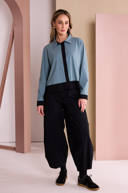 A woman modeling a two-tone long-sleeve collared shirt in light blue and black, paired with black wide-leg pants and black sneakers, standing in a studio with beige and pink architectural props.