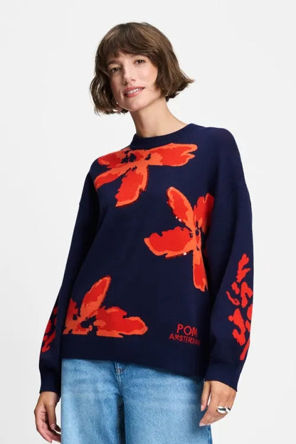A woman wearing a navy blue crewneck sweater with large red abstract floral patterns and 'POM AMSTERDAM' branding near the lower right front, paired with light blue jeans against a plain white background.