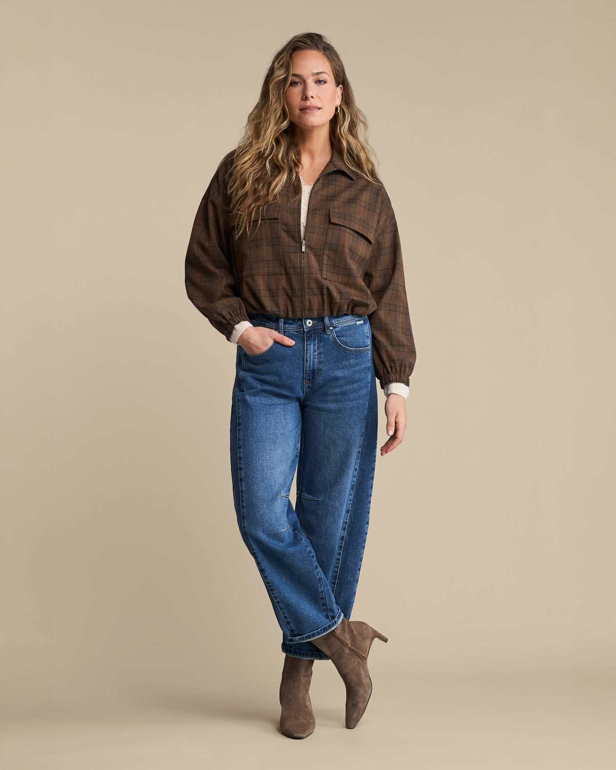 A woman modeling a brown plaid button-up shirt with a half-zip, blue jeans, and brown suede ankle boots against a solid beige studio background.