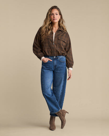 A woman modeling a brown plaid button-up shirt with a half-zip, blue jeans, and brown suede ankle boots against a solid beige studio background.