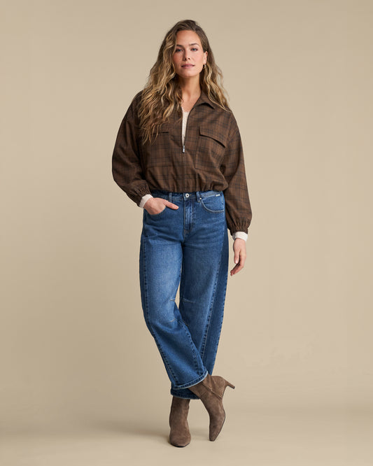 A woman modeling a brown plaid button-up shirt with a half-zip, blue jeans, and brown suede ankle boots against a solid beige studio background.