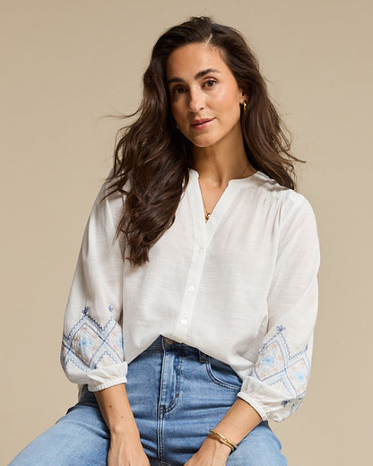 Woman wearing a white blouse with patterned sleeves and blue jeans against a beige background