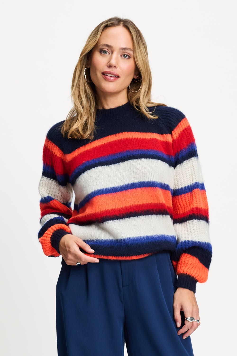 A woman wearing a fuzzy knit sweater with horizontal stripes in navy blue, red, orange, and off-white, paired with navy blue trousers, against a plain white studio background.