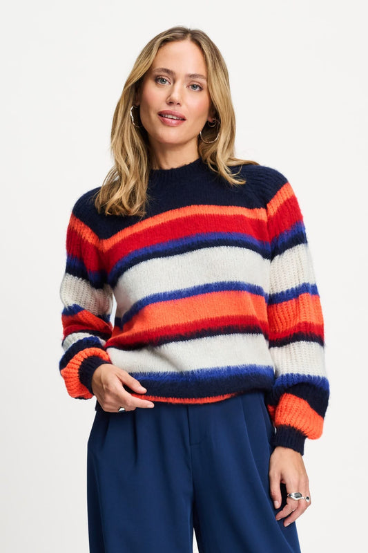 A woman wearing a fuzzy knit sweater with horizontal stripes in navy blue, red, orange, and off-white, paired with navy blue trousers, against a plain white studio background.